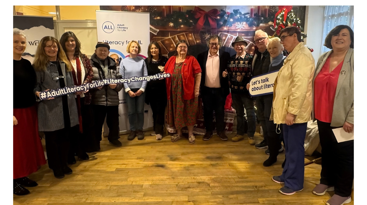 A group of people standing indoors with signs promoting adult literacy, in front of festive decorations and literacy-themed banners.